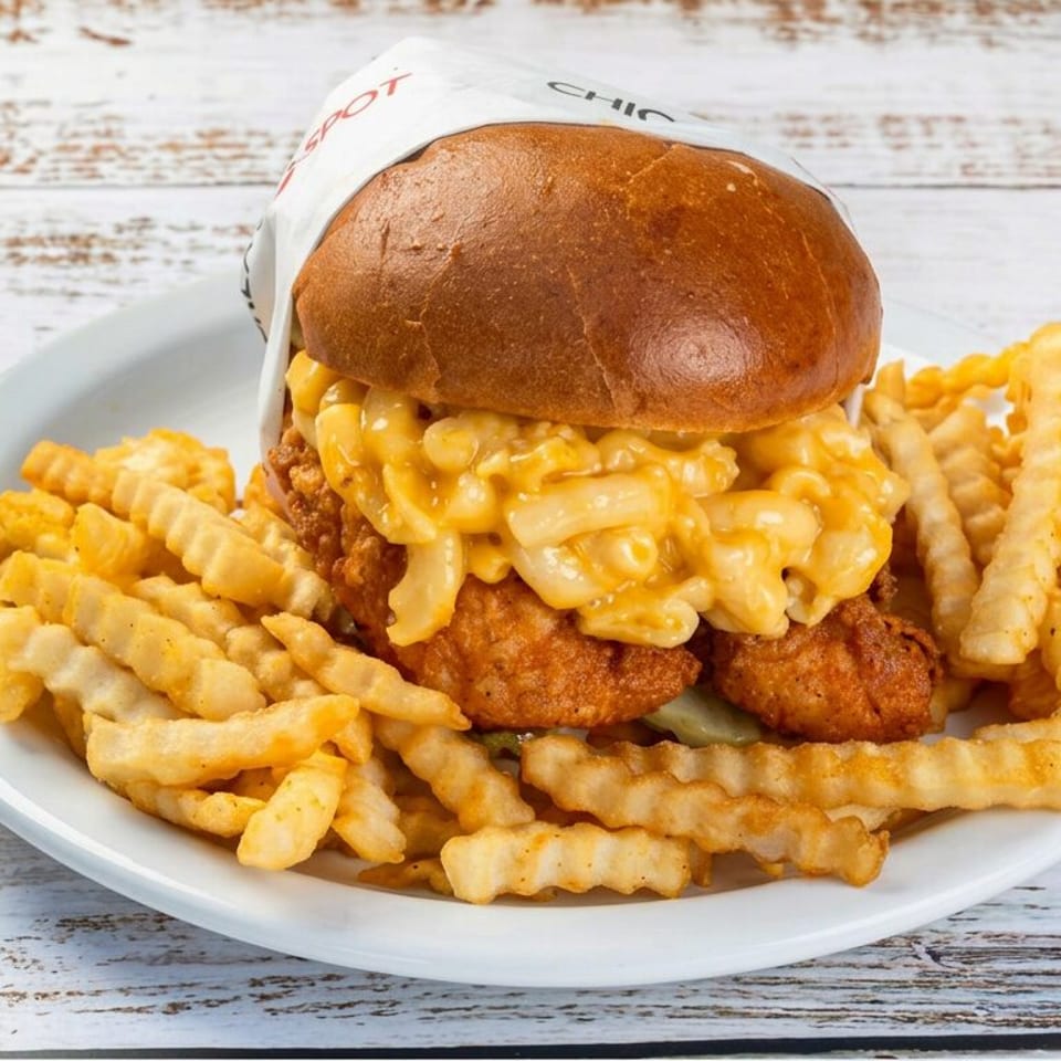 Crispy chicken sandwich topped with macaroni and cheese, served with crinkle-cut fries on a white plate.